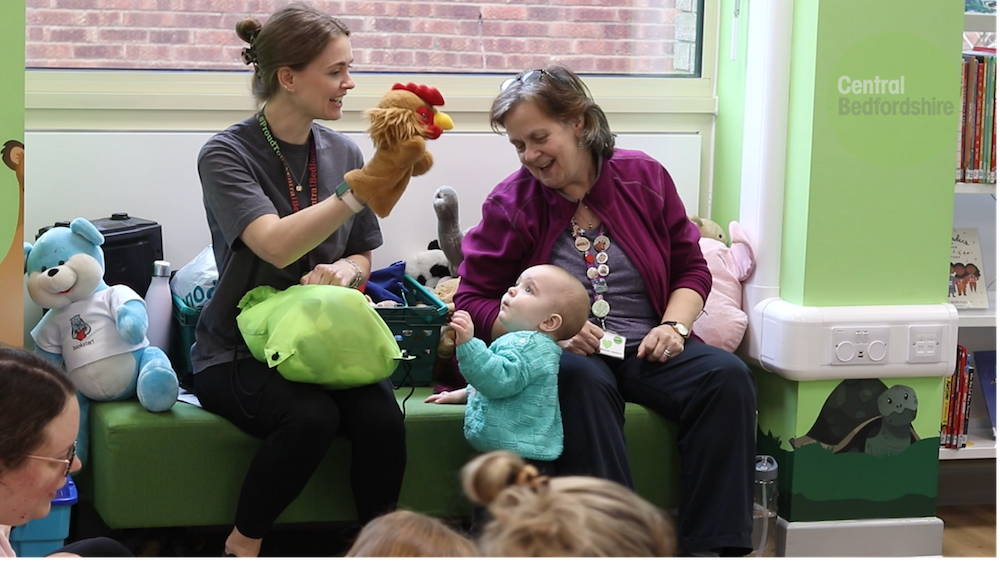 Two women teaching a music lesson. One has a puppet on her hand and a toddler is staring up at it.