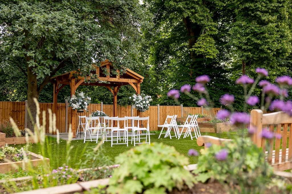The garden at the Court House, Ampthill. Lawn and colourful flower beds border on a lawn area with a natural wood gazebo with white seating.