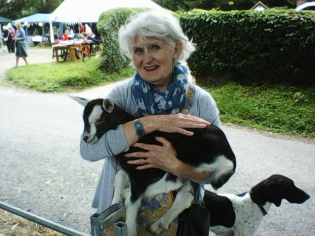 A photo of Ruth holding a baby goat. She's smiling and has a dog standing behind her.